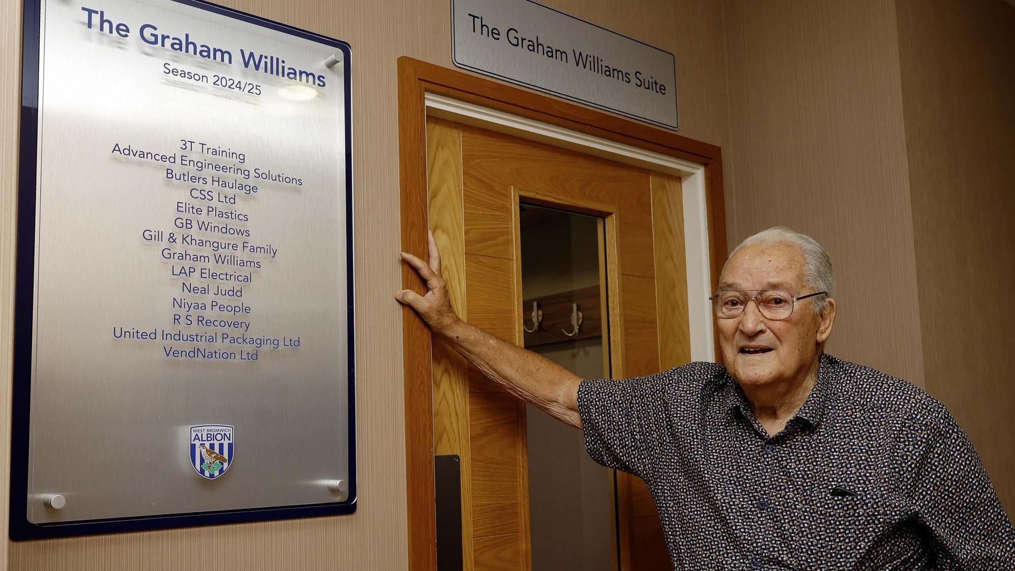 An elderly man with white hair and silver wire-framed glasses leans against a wooden internal door with a plaque outside it denoting his name and sponsors. A sign above the door reads Graham Williams suite. He is wearing a black and white short-sleeved shirt.