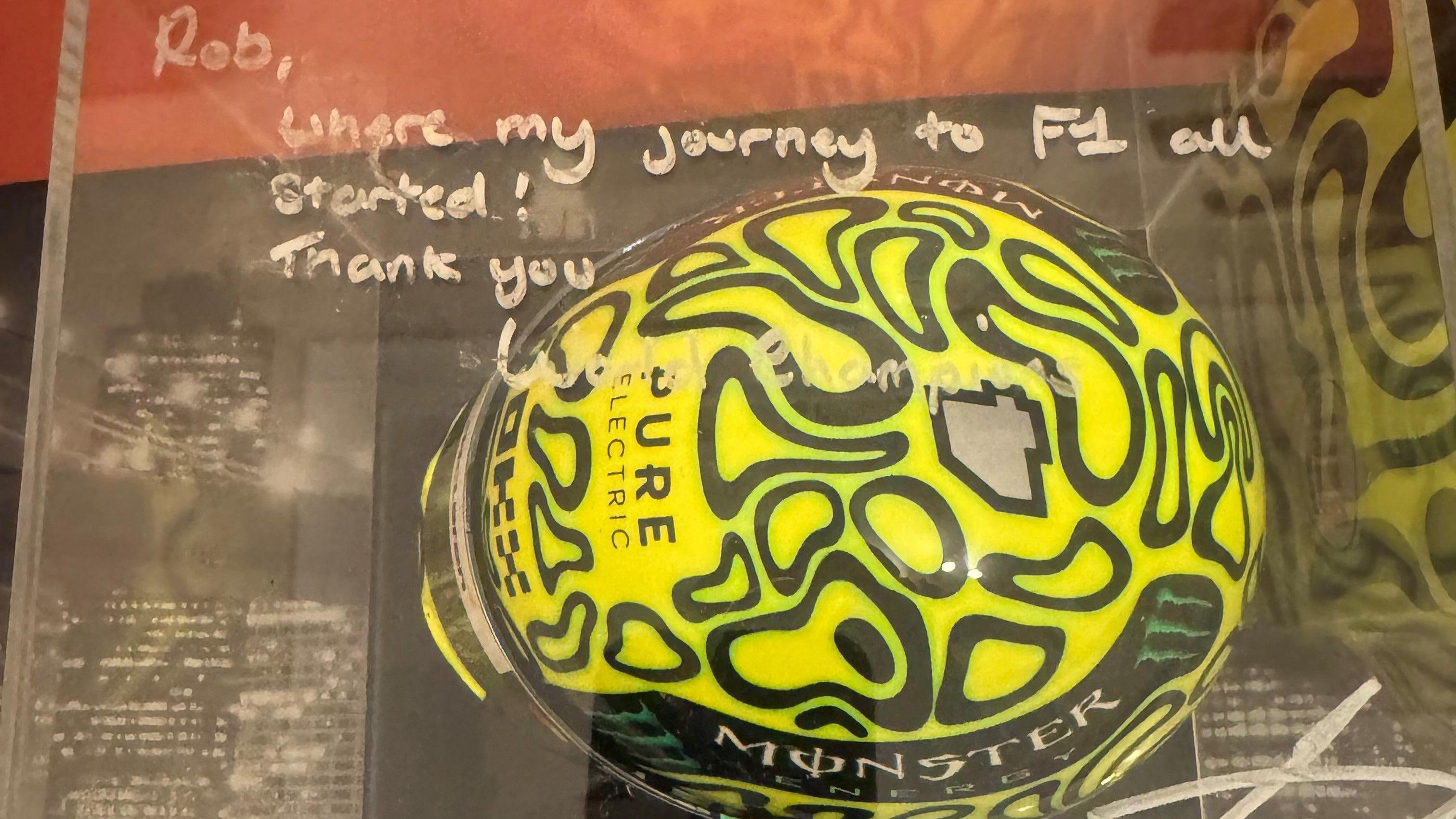 A Perspex case contains a neon yellow helmet. The helmet has a design with black shapes across it which never touch. On the surface of the frame is the message Norris left Dodds in white marker.