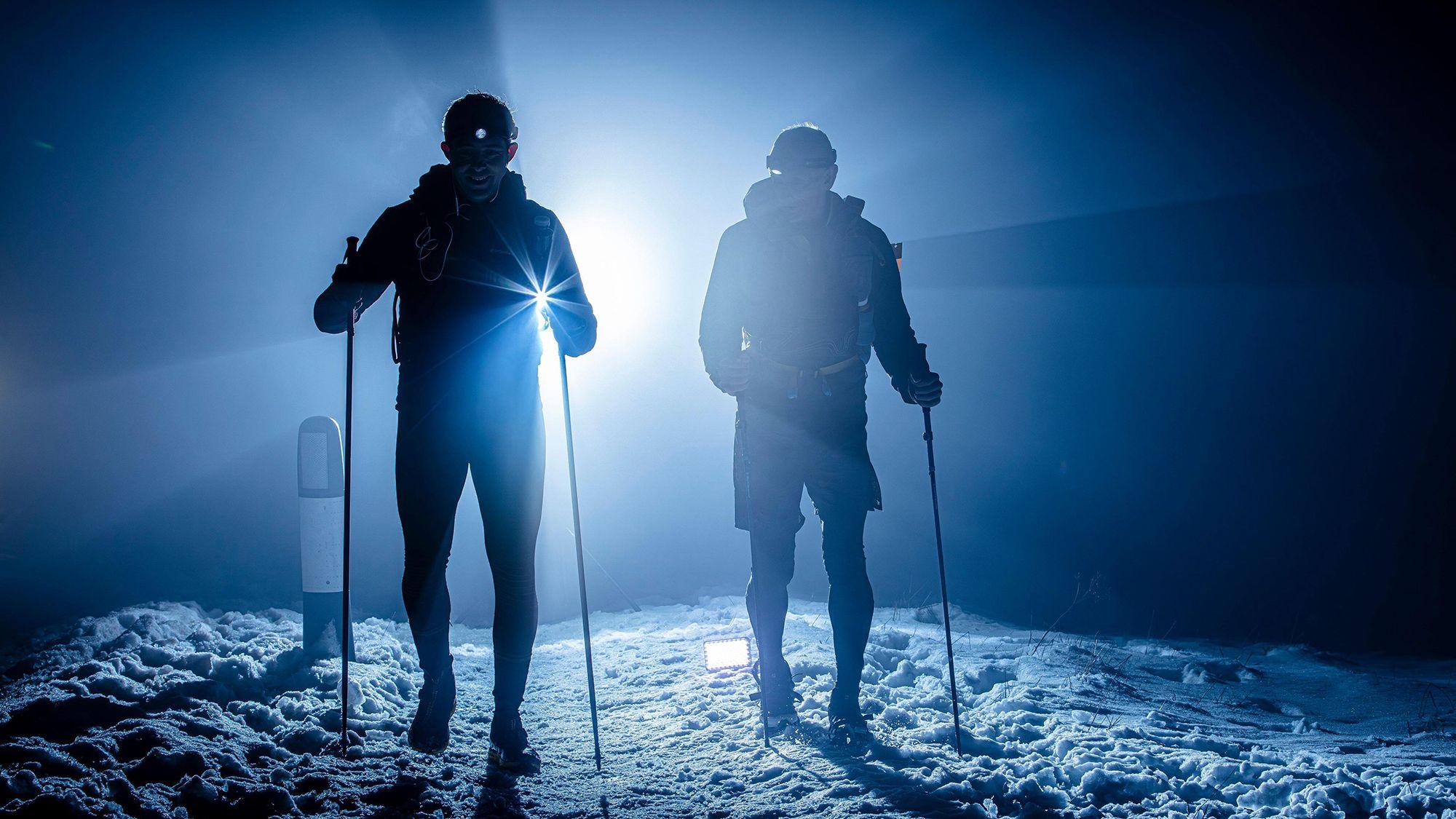 Two racers with running poles on snow, silhouetted against a bright backlight during nightfall.