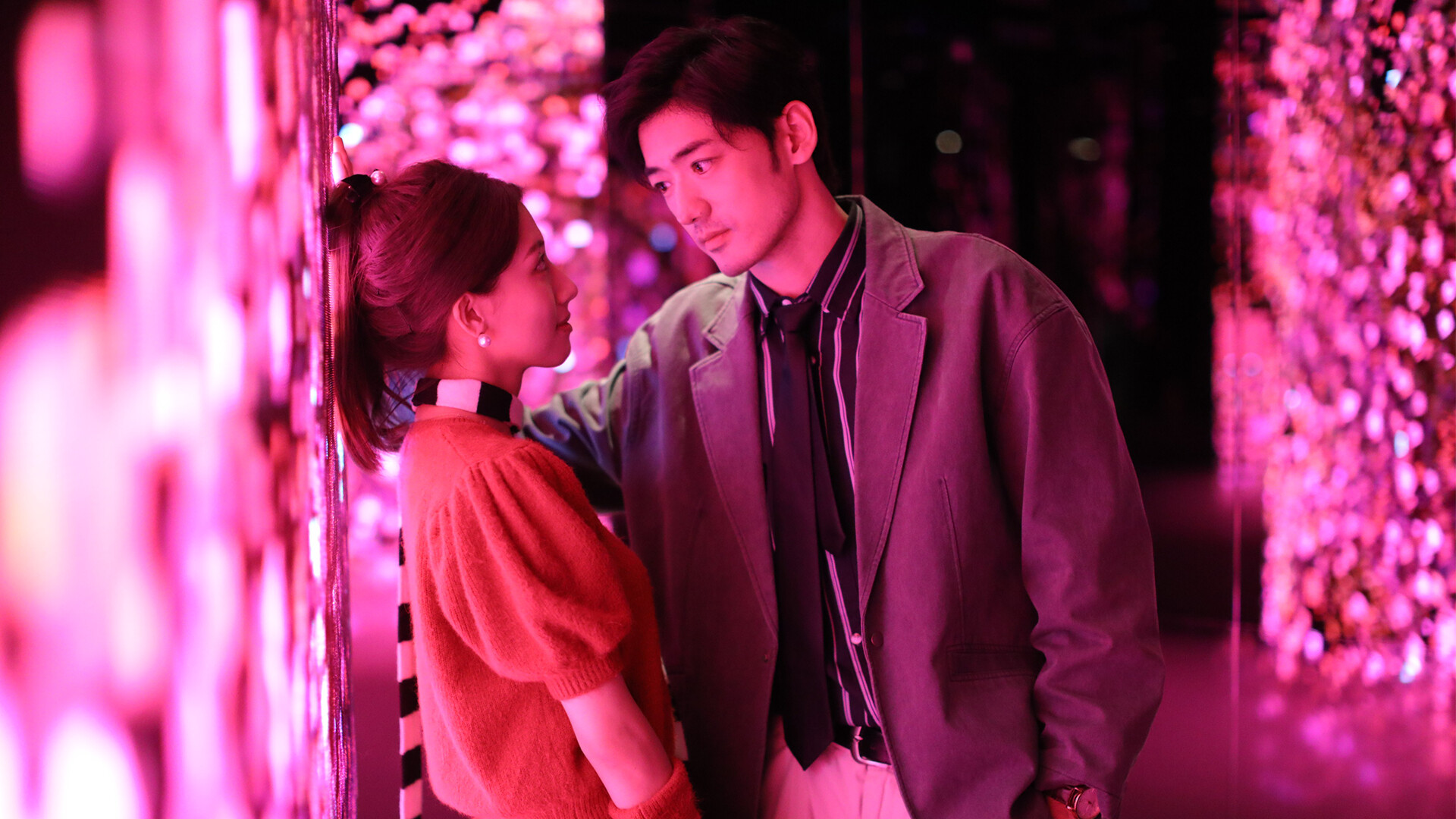 A man and a woman gaze into each other's eyes in an outdoor area bathed in pink light.
