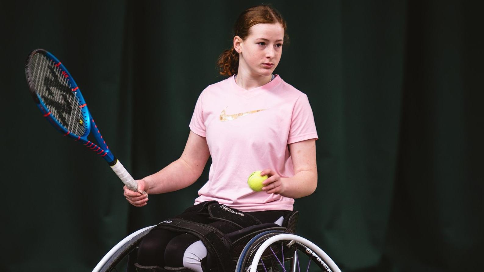 A girl in a wheelchair sits side-on while holding a tennis racket in one hand and a tennis ball in the other. She is wearing a pink T-shirt and black trousers, and has auburn hair tied back.