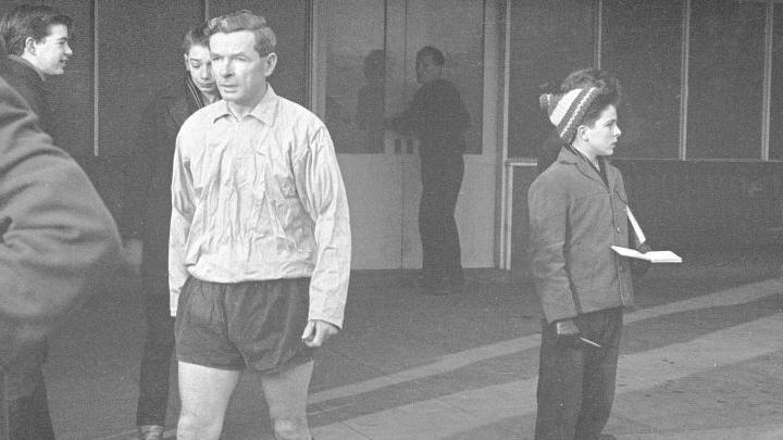 A black-and-white image of a man in a long-sleeved shirt, shorts and football socks walks along a paved area in front of a glass-fronted building. A child with an autograph book stands several paces away, looking away.