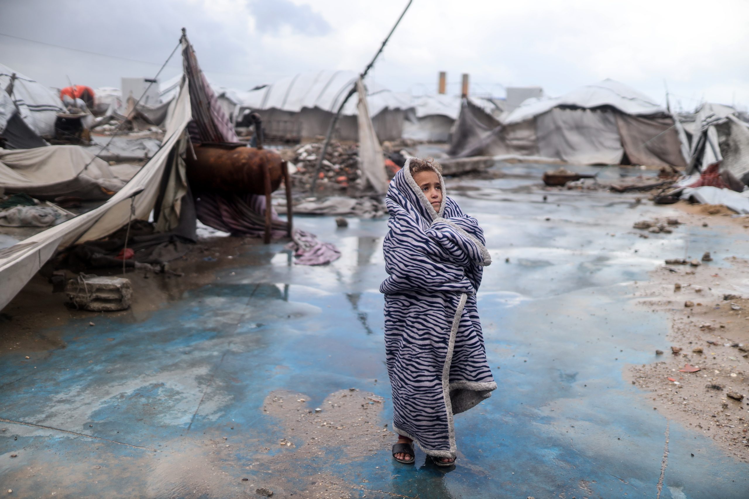 A displaced Palestinian girl shields herself from the cold as she stands in front of her family's tent after it is damaged by a storm at a displacement camp in Gaza City, Palestine, on January 13, 2026. Photo by Majdi Fathi/NurPhoto via Getty Images