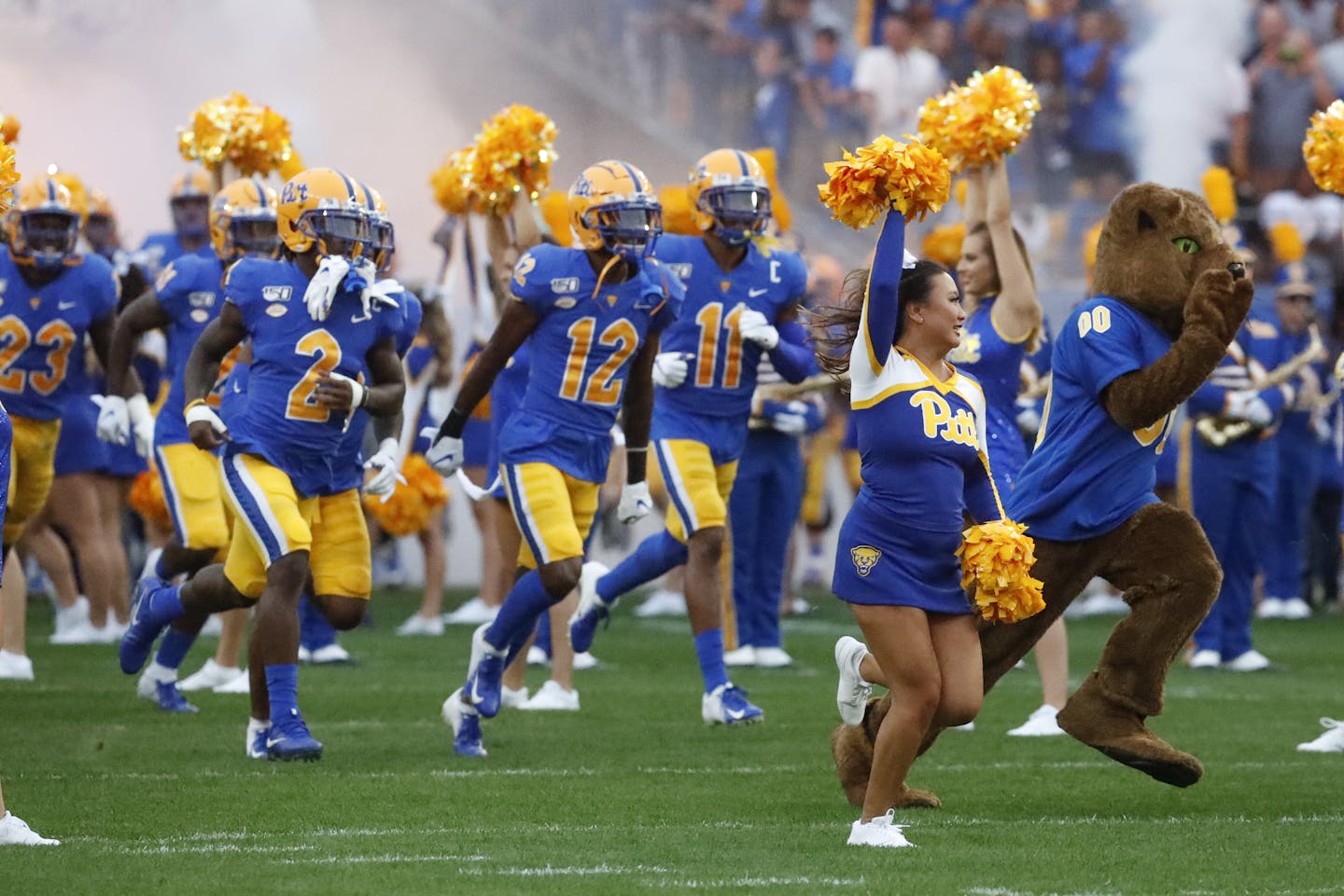 Football players and cheerleaders wearing royal blue and gold run on to a football field.