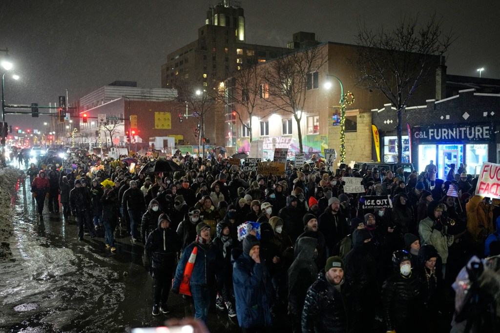 a large crowd marches down a wet city street at night during a snowstorm holding protest signs
