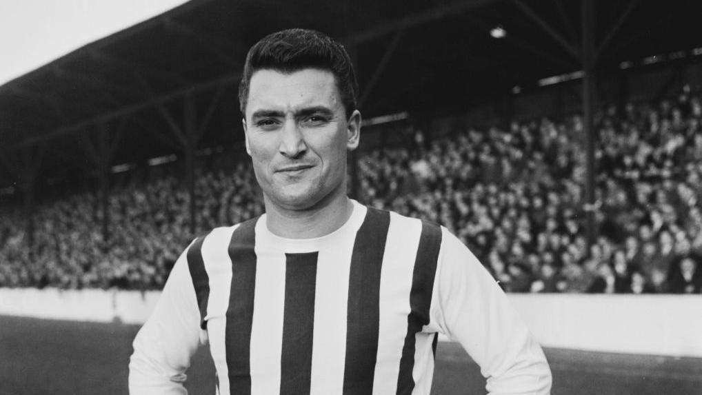 A black-and-white image of British footballer Graham Williams pictured on a football pitch with stands full of fans in the background. He is wearing a striped football jersey.