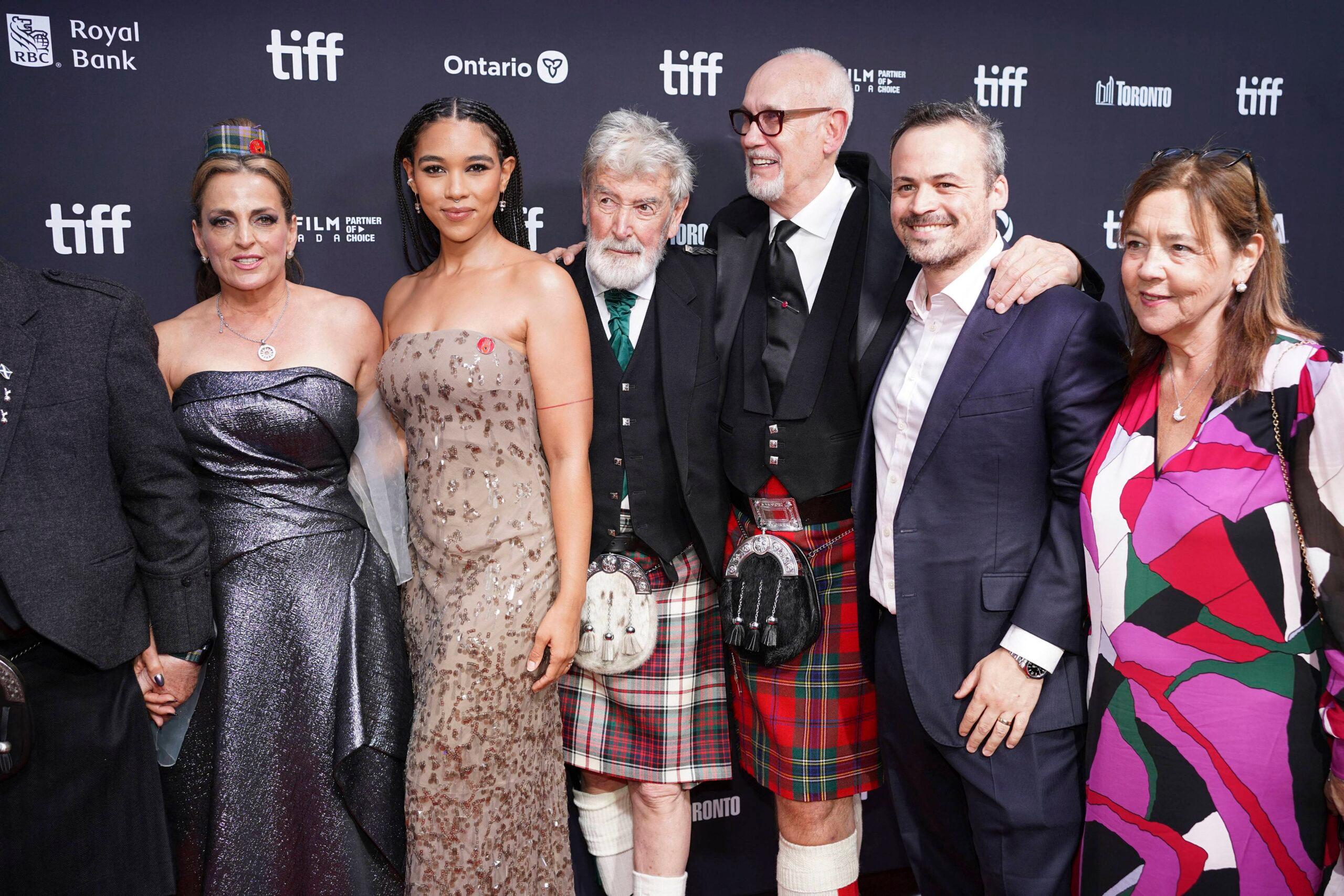 Film producer Neil Zeiger wearing a tartan kilt and glasses, surrounded by actors