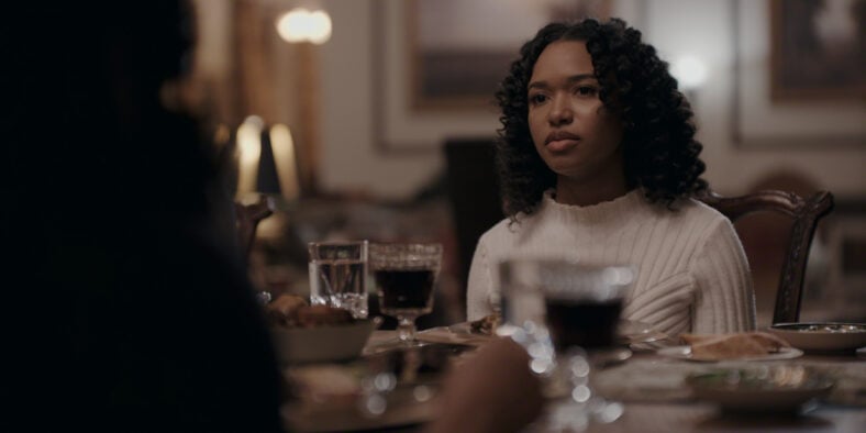 "A young woman in a white sweater sits at a dinner table set with glasses and plates, in a softly lit, elegant dining room. She looks thoughtful or serious, suggesting a quiet or tense mood."