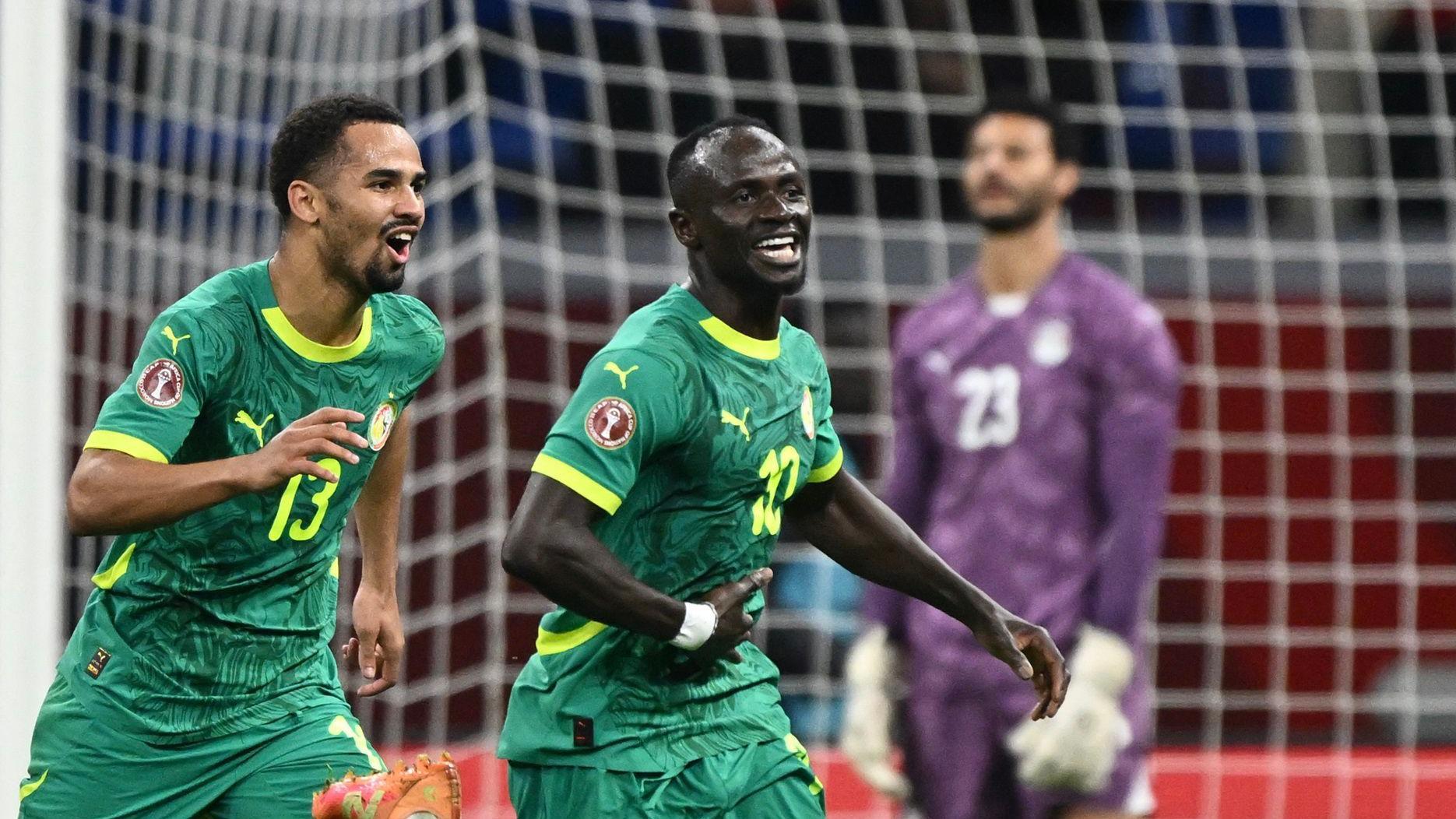 Sadio Mane wheels away in celebration with a smile on his face as he is chased by Senegal team-mate Iliman Ndiaye. Both players wear green Senegal kits, while Egypt goalkeeper Mohamed El Shenawy, wearing purple, is seen dejected in the background standing in front of a blurry white net