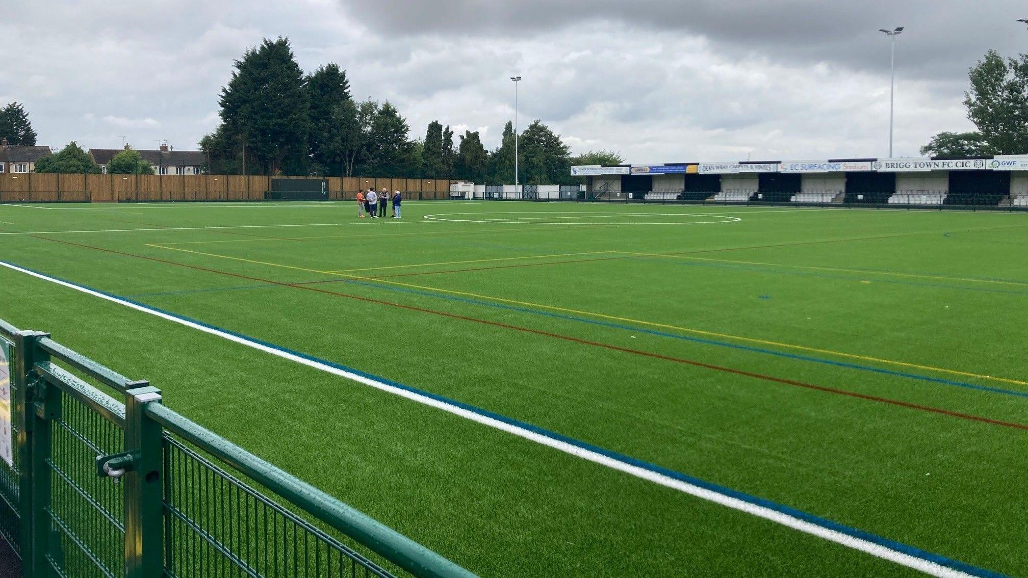 A wide view of the pitch and the stand. The surface is bright green with newly painted lines. There is a group of people standing on the pitch in the background.
