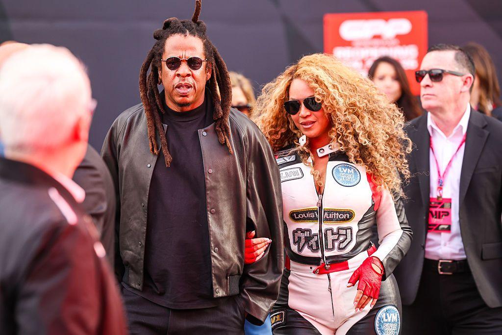 Picture of Jay-Z wearing a brown jacket with a t-shirt underneath and Beyonc&eacute; wears a white racing suit with red and black accents.