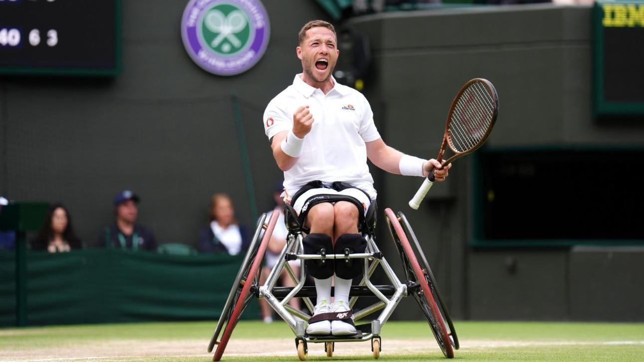 Alfie Hewett, wearing white tennis gear, pumps his right fist in triumph on the court at Wimbledon. He is in a specialist wheelchair and is holding a racket in his left hand.