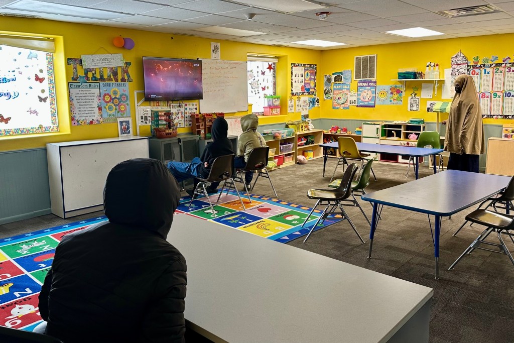 three children in hoodies watch a screen in a bright yellow classroom while a woman in a hijab stands nearby