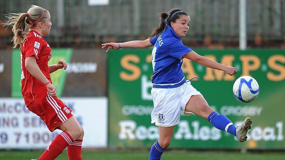 Two female football players, one with long brown hair and the other with long blonde hair, playing the sport on the pitch