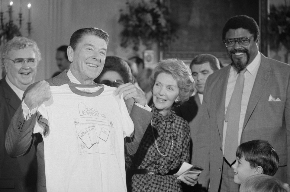 (Original Caption) Washington, D.C.: President Reagan holds up a T-shirt given him by former football star Rosie Greer promoting prayer in public schools after giving his radio address at the White House.