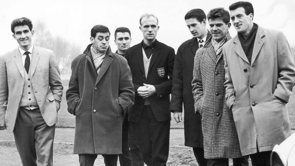 A black-and-white image of seven men, taken in 1963, walking close together. The men look as though they are in the middle of a conversation. One is looking at the photographer and smiling. All are wearing either overcoats or blazers and pullovers, with shirts and ties. They are walking away from a grassy area.